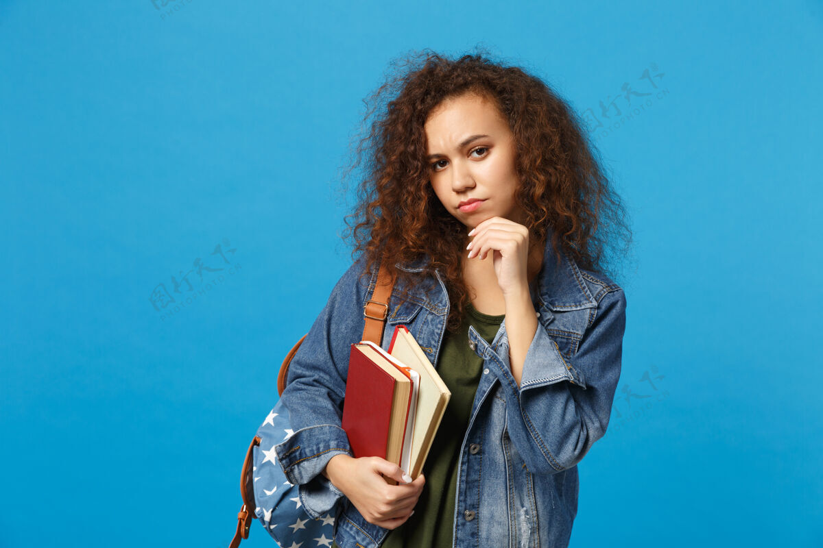 学生穿着牛仔服 背着书包的悲伤的年轻学生把书孤立地放在蓝色的墙上背包学校阅读