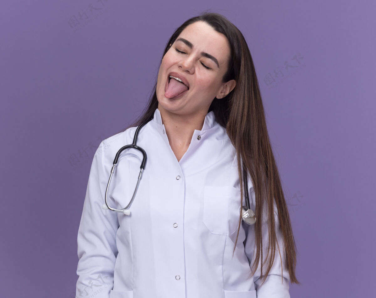 医生快乐的年轻女医生穿着带听诊器的医用长袍 把舌头堵住了医学女性长袍
