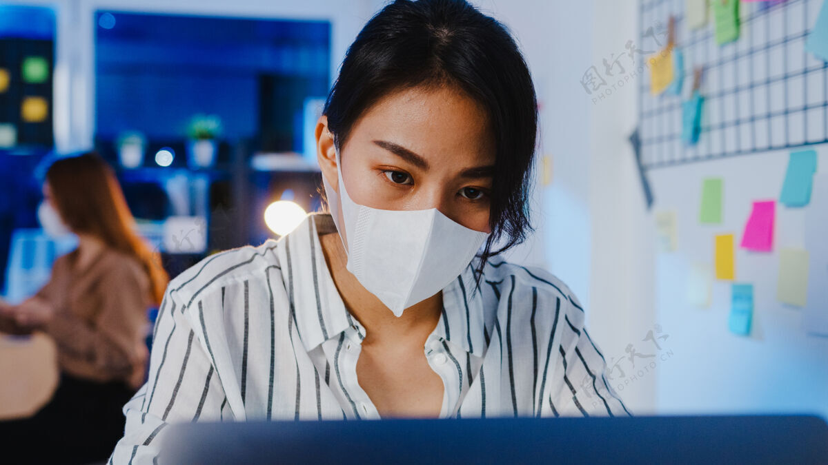 西班牙裔快乐的亚洲女商人戴着医用口罩 在新的正常情况下保持社交距离 以防病毒 晚上在办公室使用笔记本电脑迟到流行病疲劳