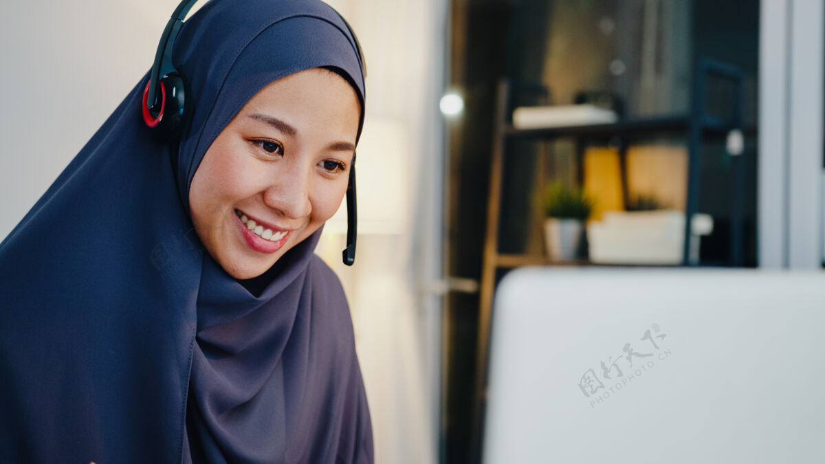 远程穆斯林女士戴着耳机看网络研讨会听在线课程在晚上的家庭办公室通过电话会议进行交流网络会议服务