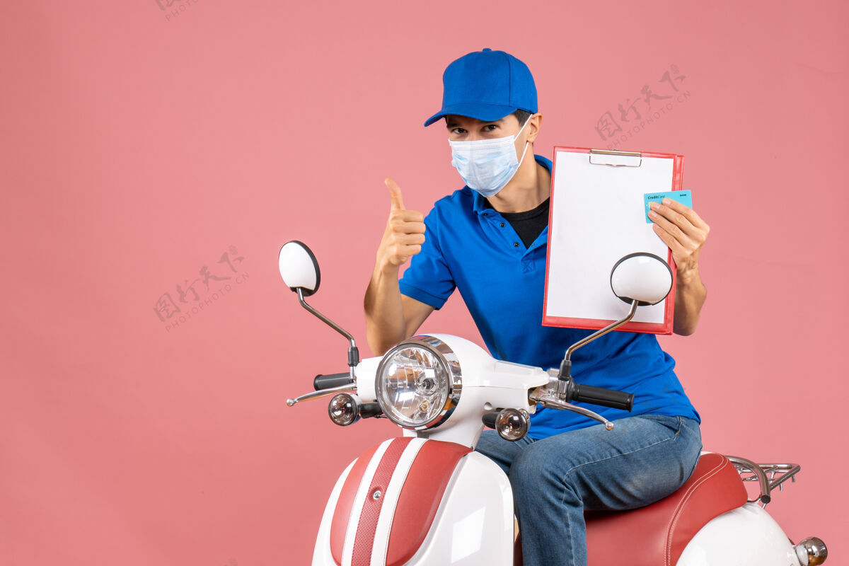 背景戴着帽子戴着面具坐在滑板车上的男性送货员的俯视图 在柔和的桃色背景上显示文件和银行卡正在做ok手势文件面具显示