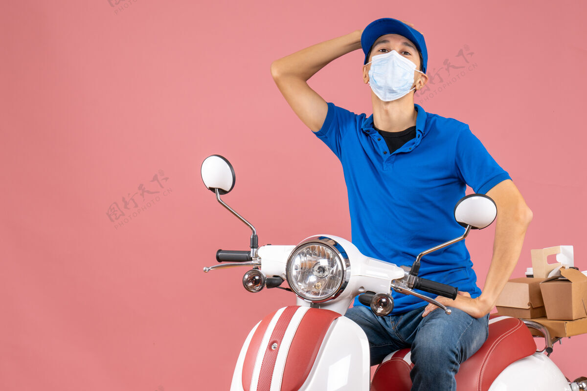 穿顶视图混乱的快递员戴着医疗面具戴着帽子坐在粉彩桃色背景的踏板车上背景面具医学