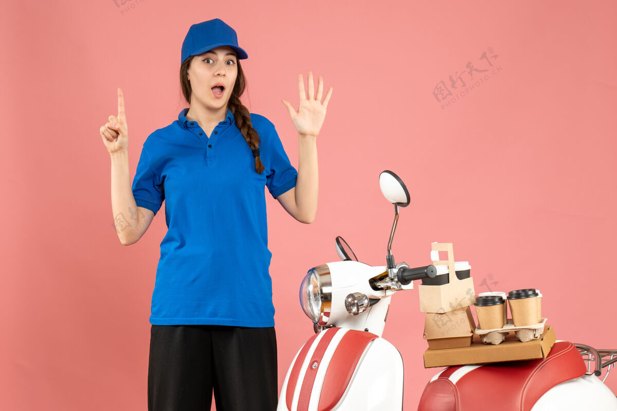 专业俯视图中的女快递员站在摩托车旁边 车上放着咖啡和小蛋糕 在柔和的桃色背景下 五个手指朝上人五咖啡