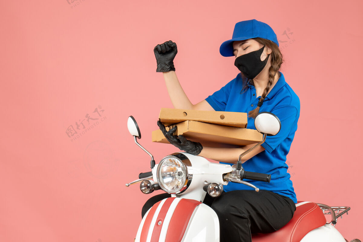 坐戴着医用面罩和手套的年轻快乐女快递员坐在粉彩桃色背景的滑板车上传递订单的正面视图穿前剪影