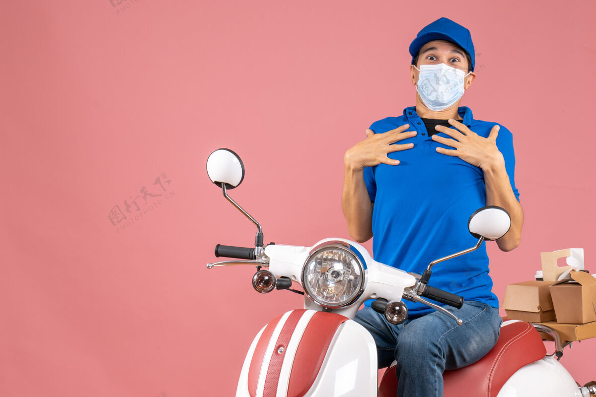 摩托车前视图不确定的快递员戴着医疗面具戴着帽子坐在滑板车上传递粉彩桃色背景的命令医疗人滑板车