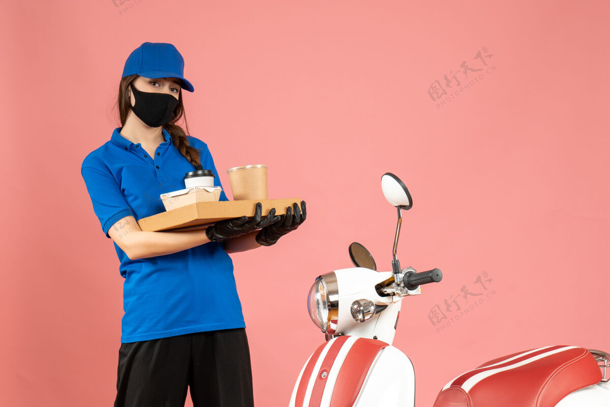 成人前视图集中快递女孩戴着医用口罩手套站在摩托车旁边拿着咖啡小蛋糕粉彩桃红色背景蛋糕咖啡手套