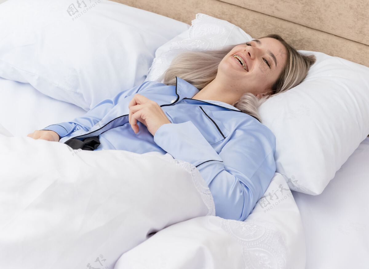 灯光穿着蓝色睡衣的年轻漂亮女人躺在床上 躺在柔软的枕头上 在卧室里享受周末的微笑背景卧室蓝色