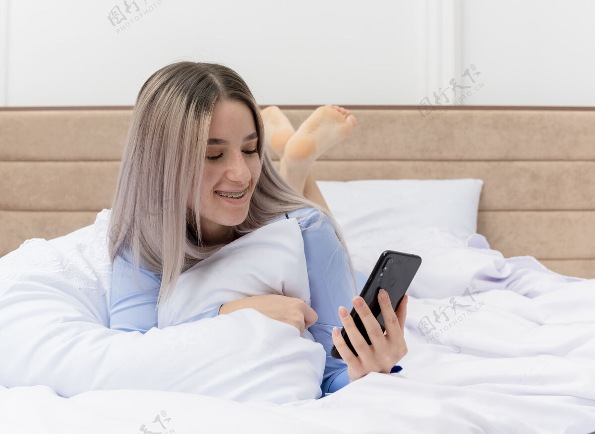 积极穿着蓝色睡衣的年轻美女躺在床上用智能手机快乐而积极地在卧室里休息快乐年轻使用