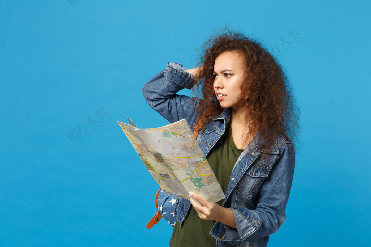 航行年轻的非洲裔美国女孩十几岁的学生在牛仔服装 背包举行地图隔离在蓝色的墙上旅游包旅行