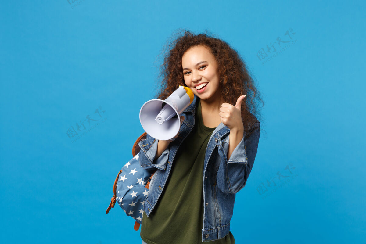 扬声器年轻的非洲裔美国少女 穿着牛仔服 背着背包 把扩音器孤立地放在蓝色的墙上包醒着人