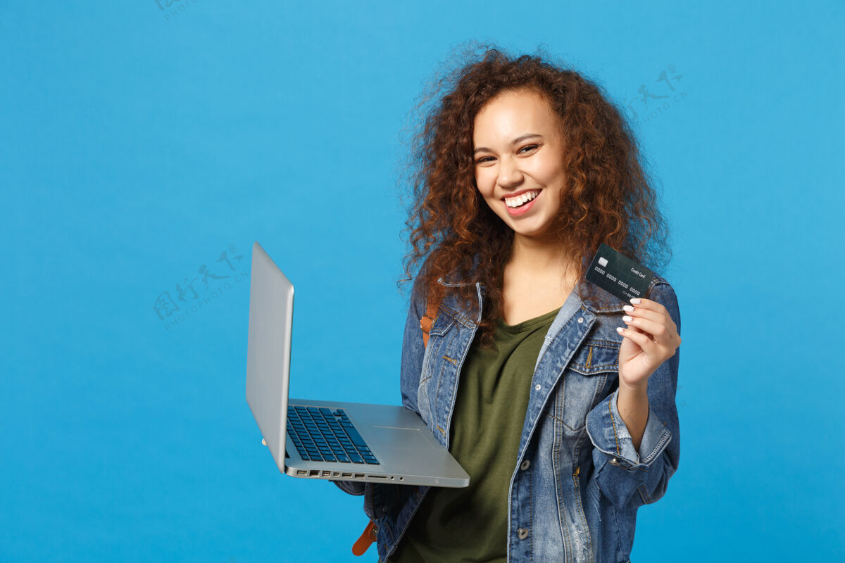 信贷年轻的非洲裔美国女孩十几岁的学生在牛仔服装 背包工作的电脑 持有银行卡隔离在蓝色的墙壁上贷款使用笔记本