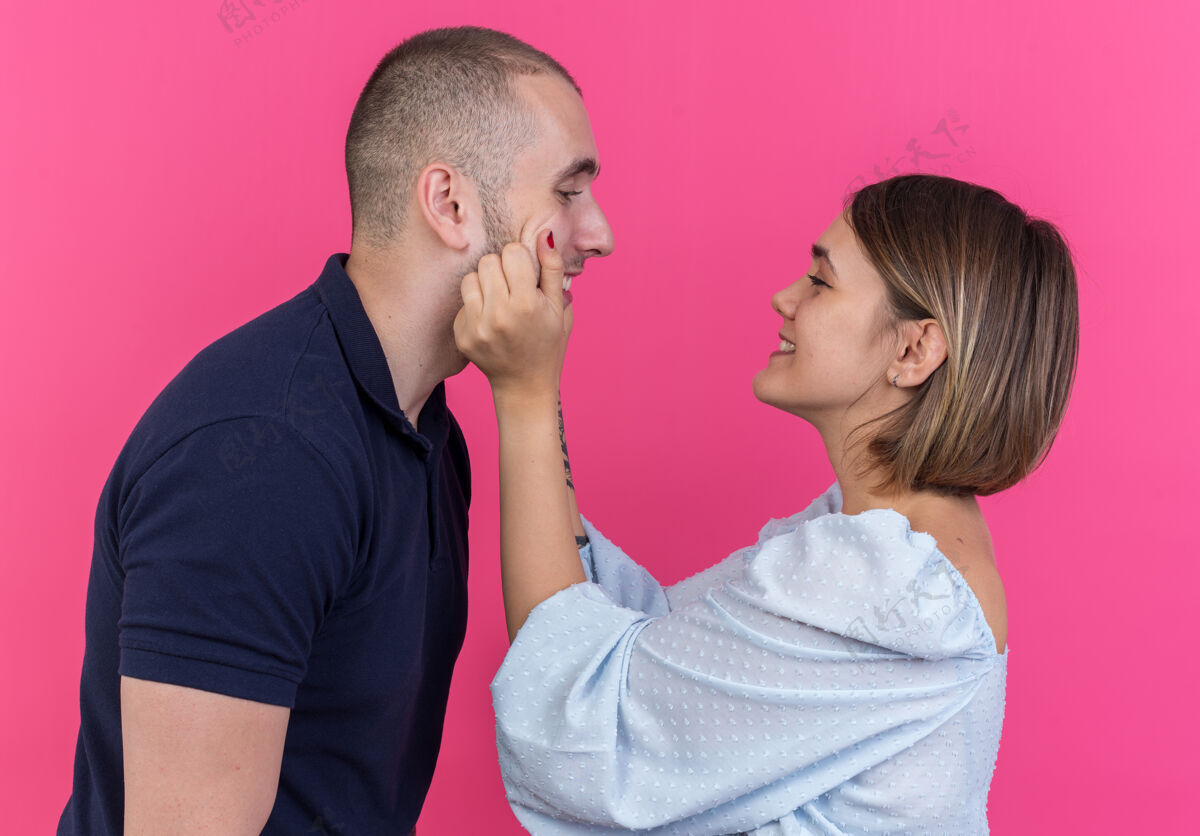 姿势快乐的年轻漂亮的夫妇微笑着的女人捏着她可爱的男朋友的脸颊站在粉红色的墙上站捏人