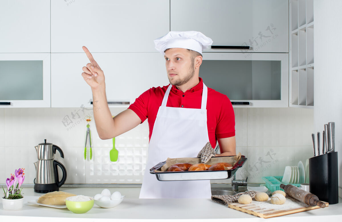指点年轻的男厨师戴着支架 拿着新鲜出炉的糕点 指着白色厨房的正视图男男厨师厨师