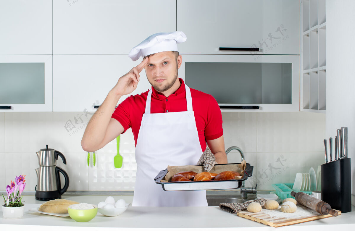 男人年轻的男厨师戴着支架手持新鲜出炉的糕点 在白色的厨房里感到自豪家庭雇员成人