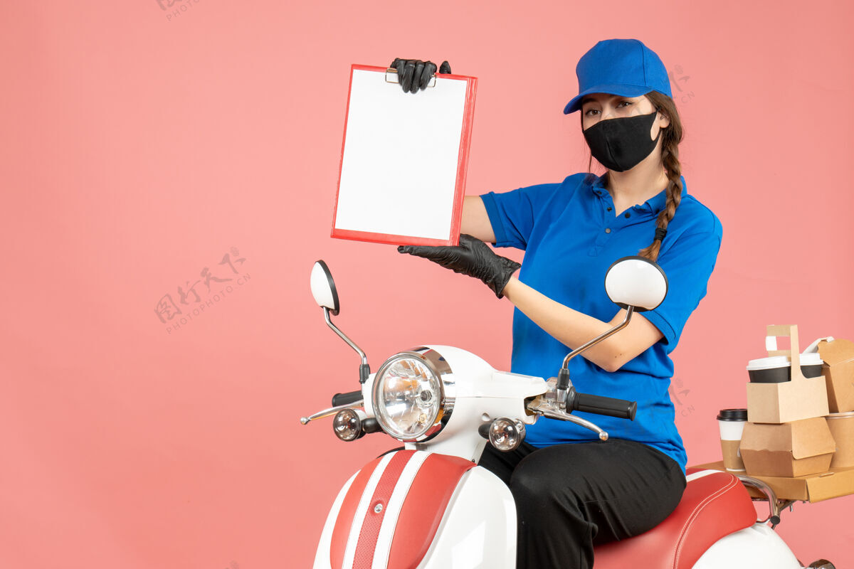 桃子前视图自信的女快递员戴着医用口罩和手套坐在踏板车上 拿着空纸 在柔和的桃色背景下传递订单粉彩面具医疗