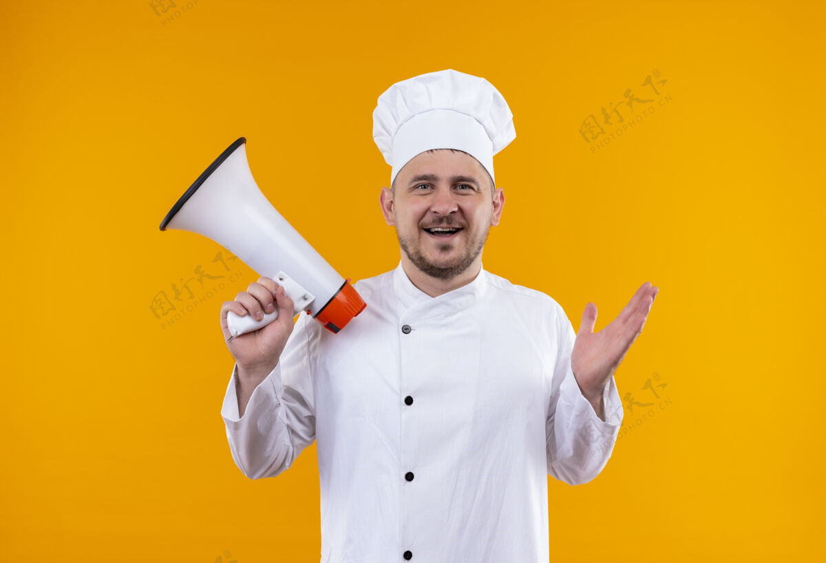 制服快乐的年轻帅哥厨师穿着厨师制服 拿着隔离在橙色墙上的扬声器烹饪演讲者厨师