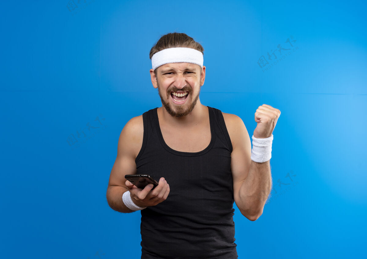 年轻快乐的年轻帅气的运动型男人戴着头带和腕带拿着手机握紧拳头隔离在蓝色的墙壁上 留着复制空间男人握紧手机