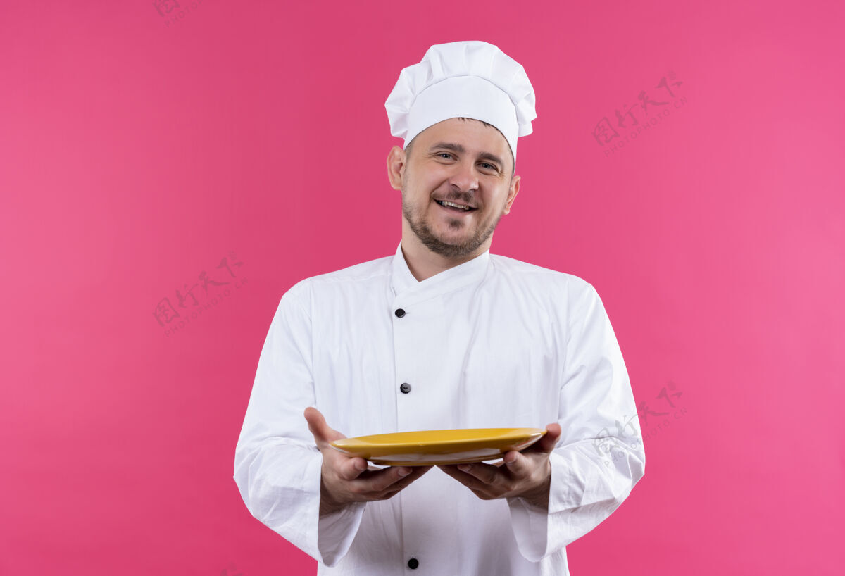 舒展快乐的年轻帅哥厨师穿着厨师制服 把空盘子摊在粉红色的墙上烹饪帅气盘子