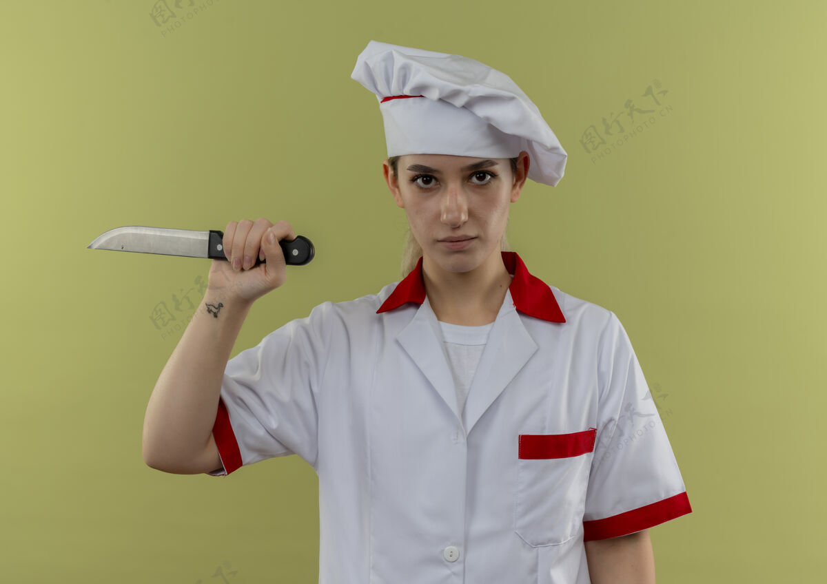 刀自信的年轻漂亮厨师穿着厨师制服 拿着隔离在绿墙上的刀持枪烹饪厨师
