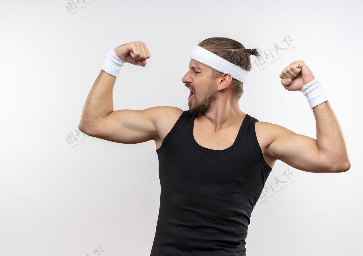 腕带自信的年轻帅气的运动型男人戴着头带和腕带做着强壮的手势 看着隔离在白墙上的肌肉强壮运动年轻