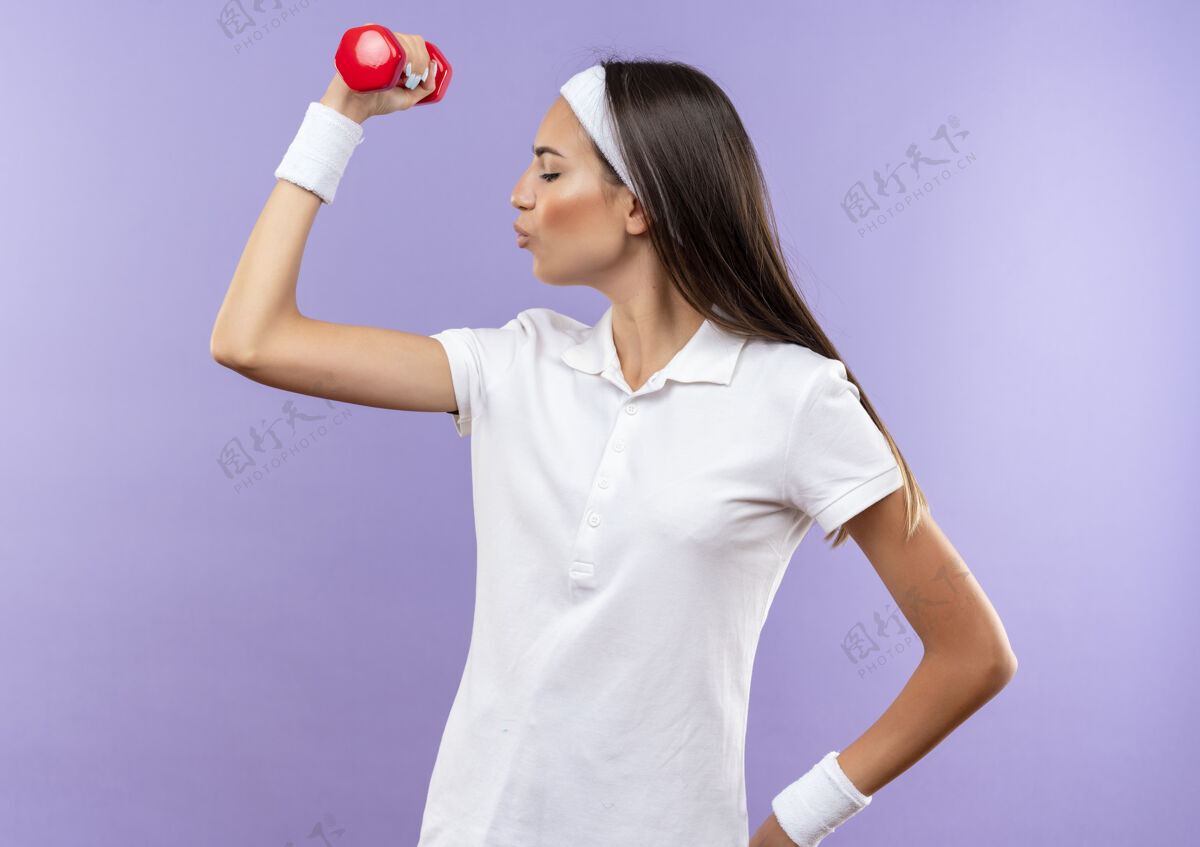 提高自信漂亮的运动女孩戴着头带和腕带举起哑铃孤立在紫色的墙上运动漂亮头带