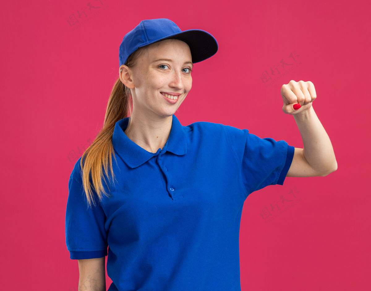 信心身穿蓝色制服 头戴鸭舌帽的年轻送货员微笑着自信 快乐 积极地站在粉红色的墙上挥舞着拳头积极交货女孩