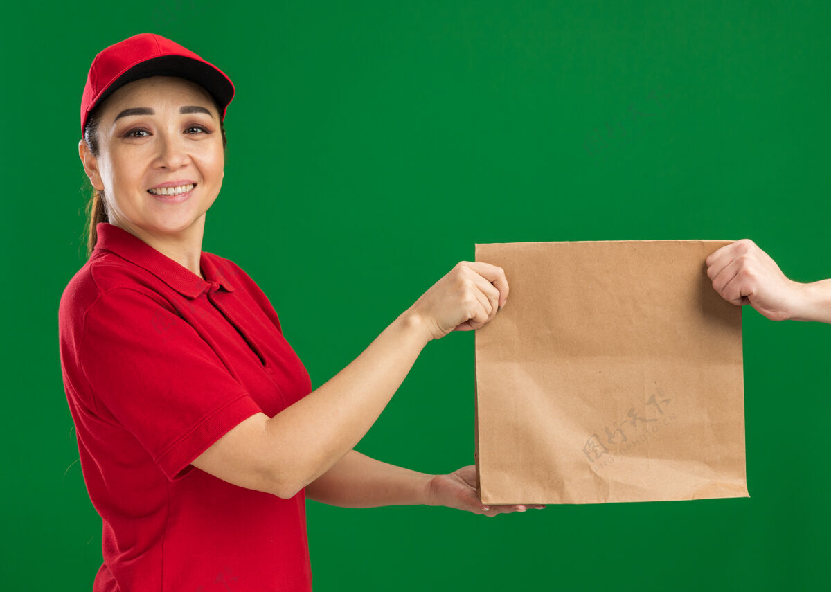 给予年轻的送货员身着红色制服 头戴鸭舌帽 站在绿色的墙边 微笑着自信地给顾客送纸包包装年轻制服