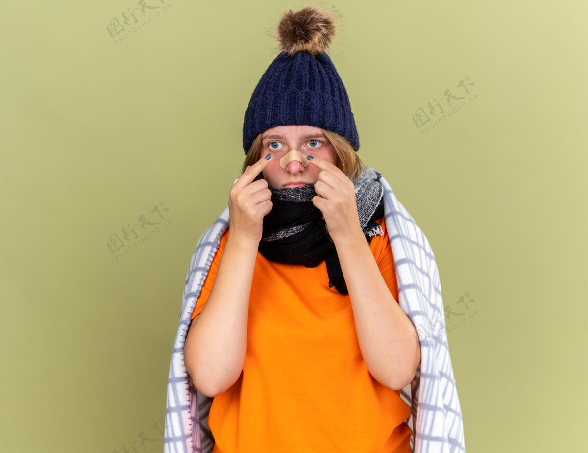 困惑不健康的年轻女子 戴着暖和的帽子 脖子上围着围巾 裹着毯子 感觉不舒服 拿着一块贴片贴在鼻子上 站在绿色的墙壁上显得困惑和担心抱着不适担心