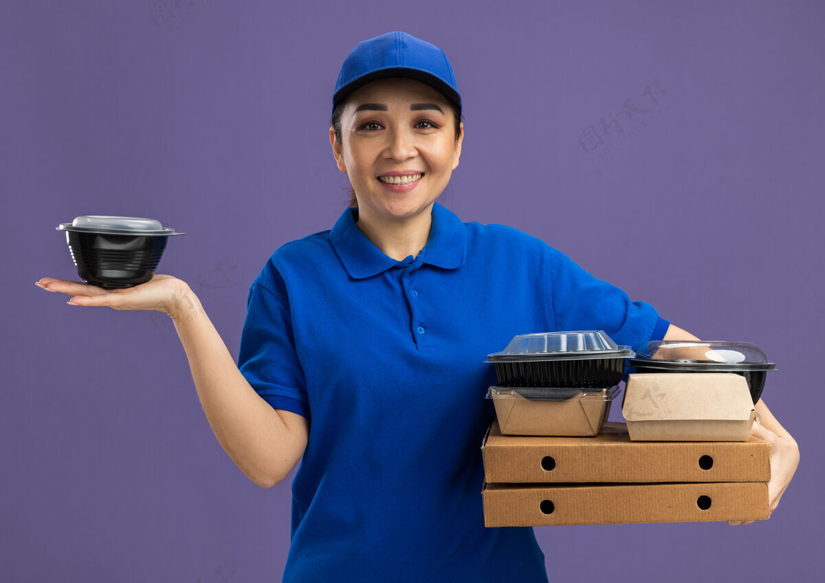 食物身着蓝色制服 头戴鸭舌帽 手持披萨盒和食品包的快乐年轻送货员站在紫色的墙上 开心地微笑着欢呼年轻人站着