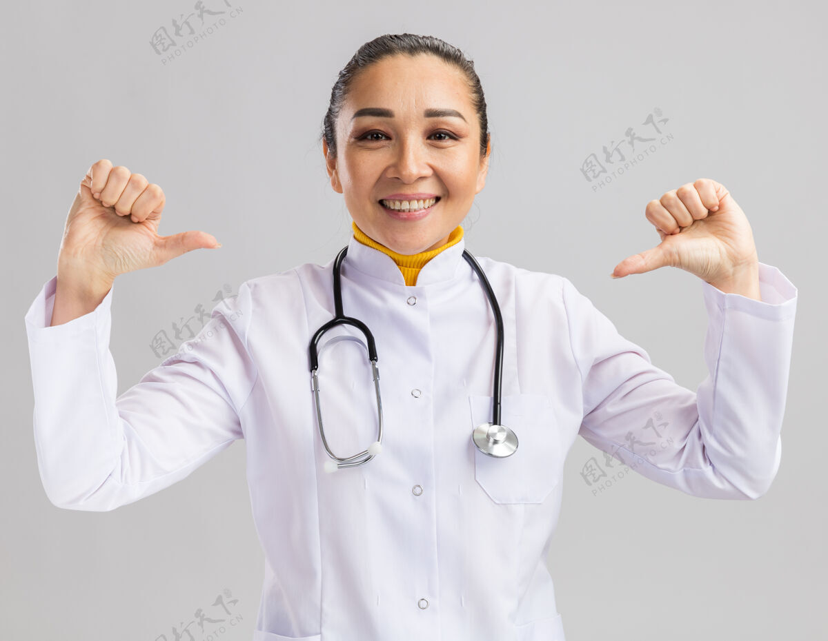 指点年轻的女医生穿着白色的医用外套 脖子上戴着听诊器 自信地微笑着指着站在白墙上的自己听诊器外套自信