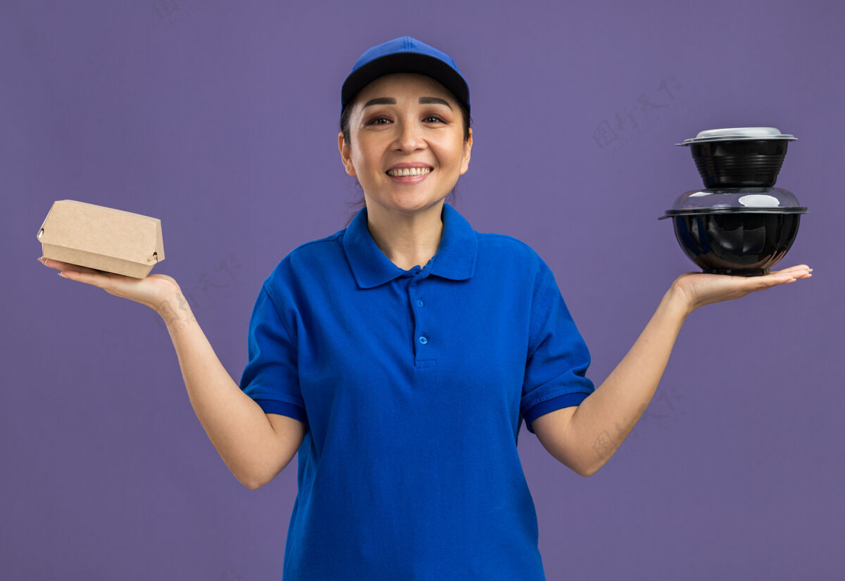 制服身着蓝色制服 戴着帽子 手里拿着食品包 站在紫色的墙上 快乐地微笑着的年轻送货员欢呼帽子年轻