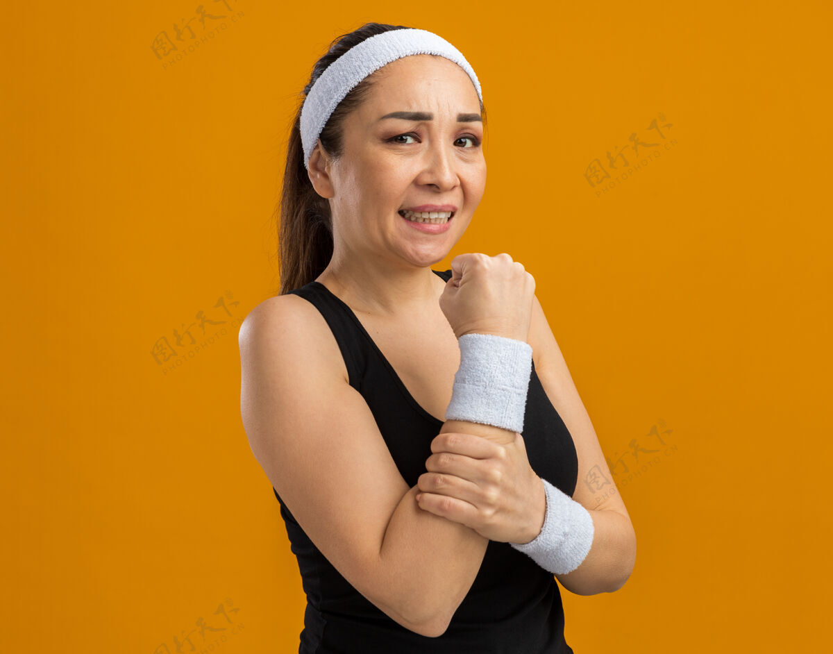 触摸戴着头带和臂章的年轻健身女士看起来身体不适 站在橘色的墙上摸着她的胳膊感到疼痛女人健康站立