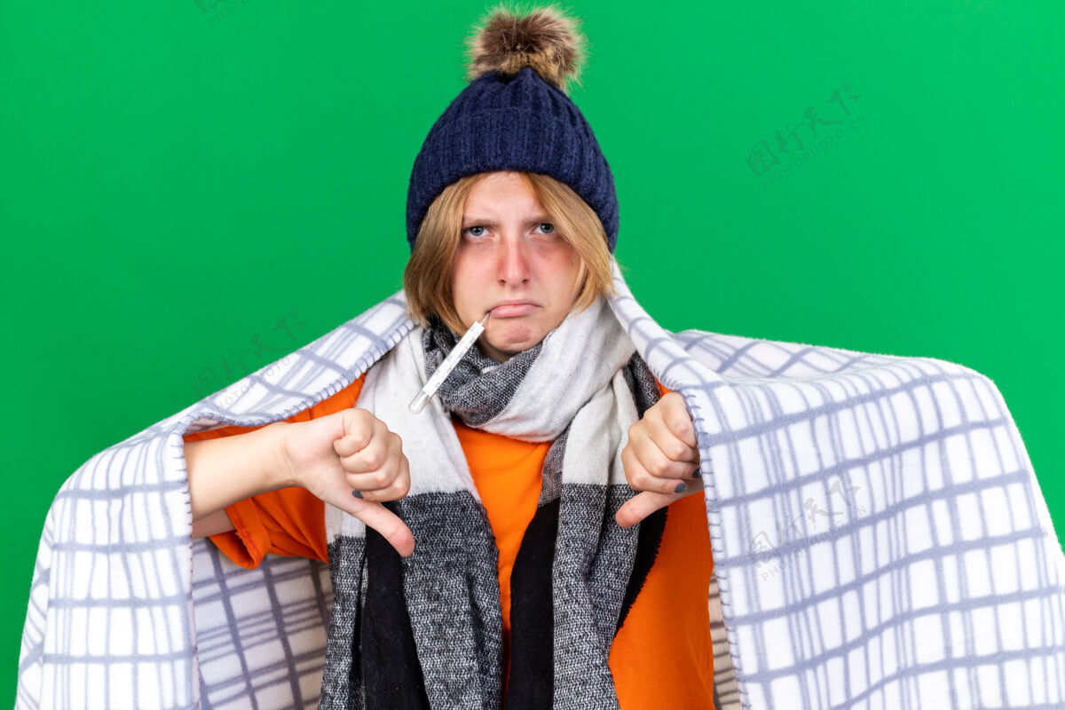包裹不健康的年轻女子裹着毛毯戴着帽子用体温计测量体温患流感发烧大拇指朝下站在绿色的墙上毯子帽子倒下