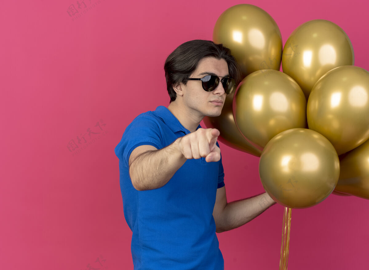 指向自信英俊的白种人戴着太阳眼镜拿着氦气球指着相机粉色氦自信