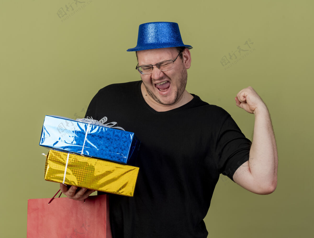 欢乐快乐的成年斯拉夫人戴着眼镜 戴着蓝色派对帽 手拿礼品盒和纸购物袋聚会空间男士