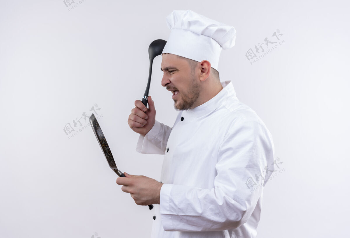 勺子愤怒的年轻帅哥厨师穿着厨师制服拿着煎锅和勺子看着孤零零的白墙上的锅帅哥年轻锅