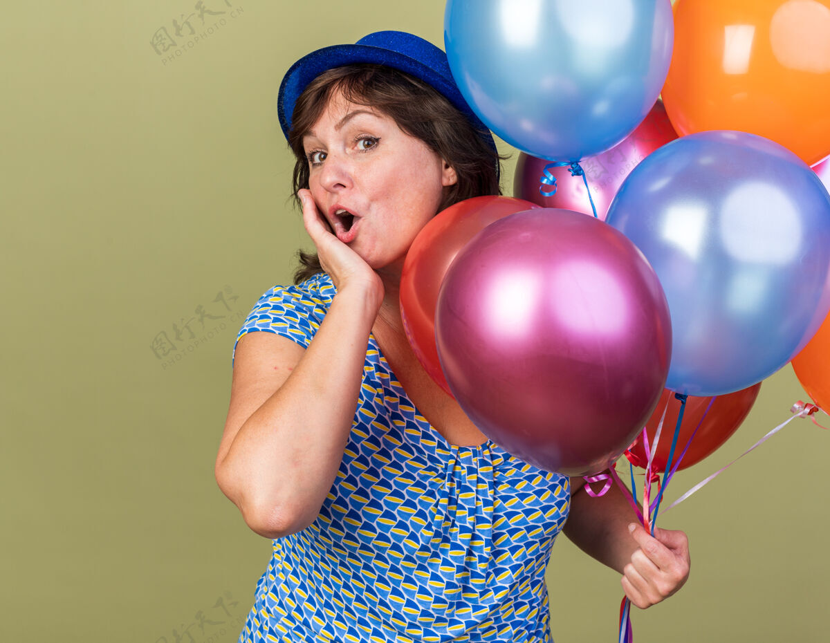 五颜六色中年妇女戴着派对帽 手里拿着一堆五颜六色的气球 站在绿色的墙上 惊喜而快乐地庆祝生日派对年龄气球立场