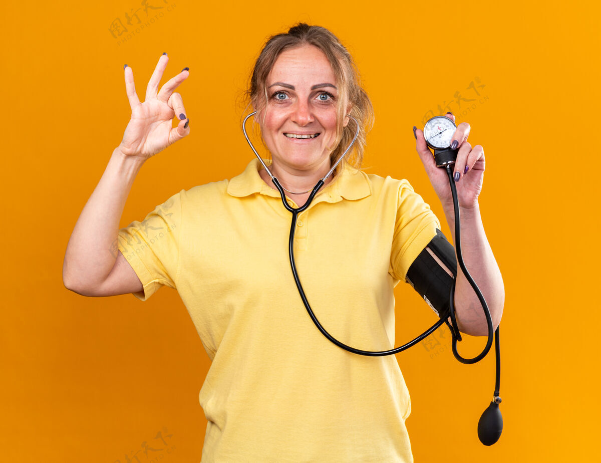 微笑穿着黄色衬衫的不健康女性感觉更好用眼压计测量她的血压微笑着站在橙色的墙上显示ok标志站着可以女人