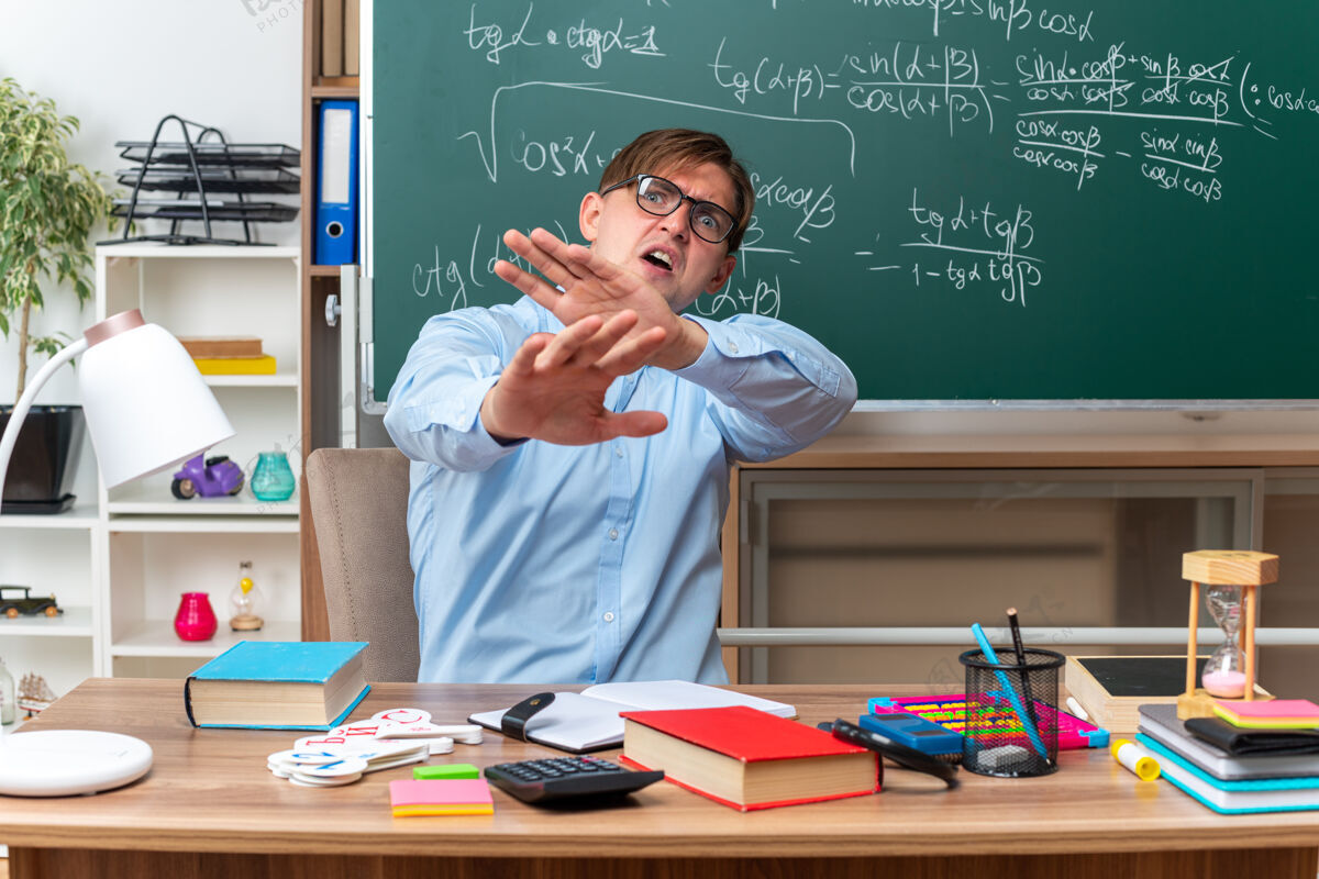 防御年轻的男老师戴着眼镜 神情焦虑 困惑地做着防御的手势 手放在课桌旁 课本和笔记放在教室的黑板前男笔记坐着