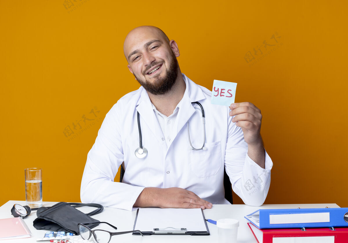 坐着高兴的年轻秃头男医生穿着医用长袍和听诊器坐在办公桌旁听诊器医疗橙色