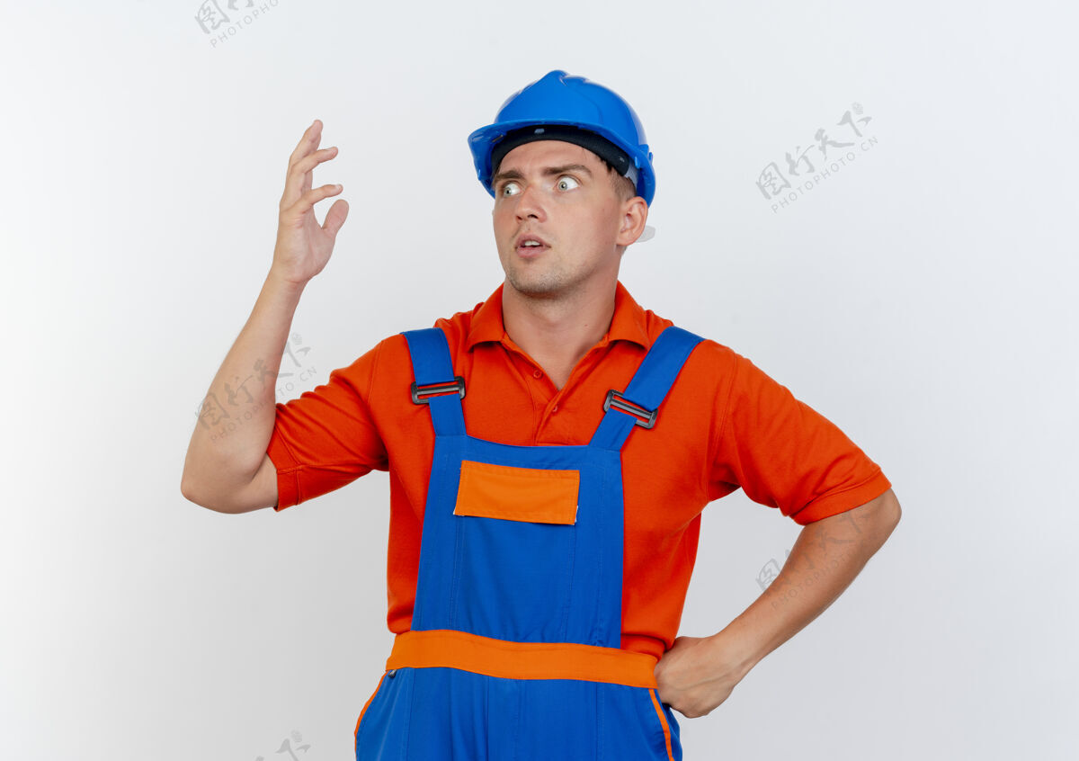 头盔看着侧面印象深刻的年轻男性建筑工人穿着制服和安全帽举起手放在臀部另一只手年轻男性佩戴