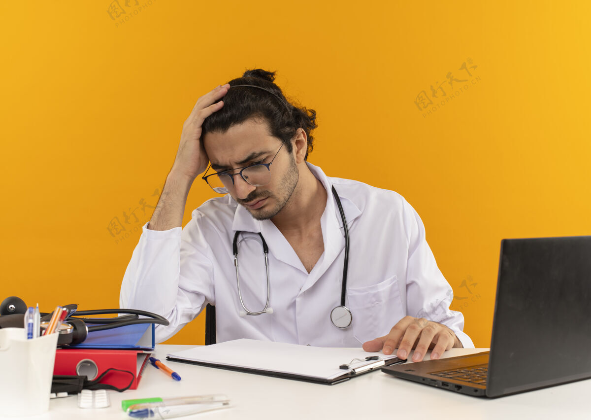 眼镜困惑的年轻男医生戴着医用眼镜 穿着医用长袍 听诊器坐在办公桌旁桌子年轻穿