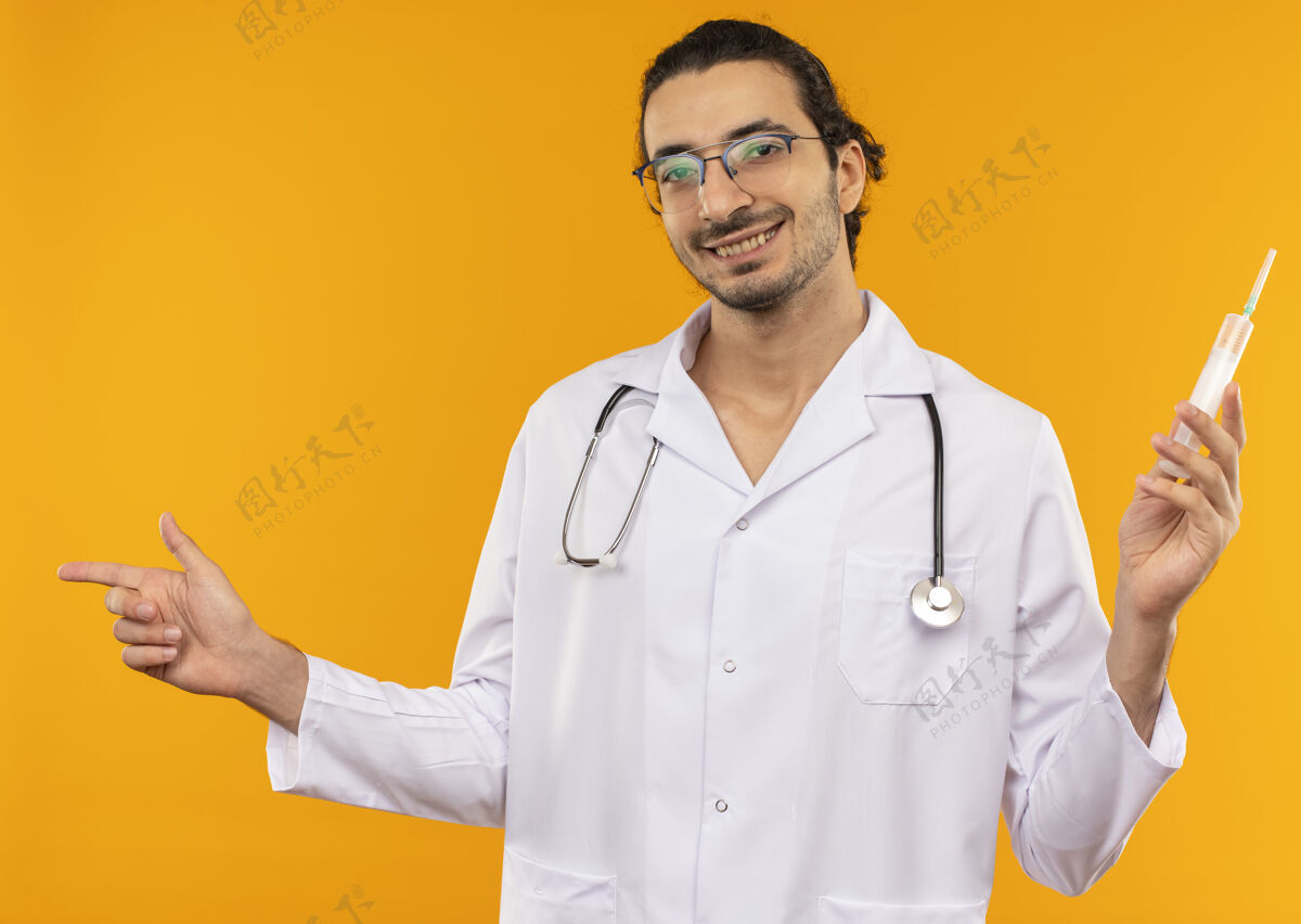 听诊器面带微笑的年轻医生戴着医用眼镜 穿着医用长袍 听诊器拿着注射器 指着旁边长袍注射器年轻