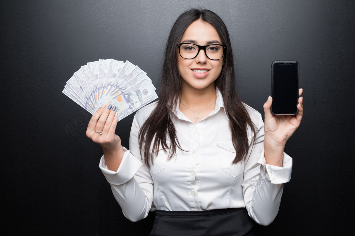 女士穿着白衬衫的快乐的年轻黑发女人展示着智能手机 黑屏上 手里拿着现金成人女人现金