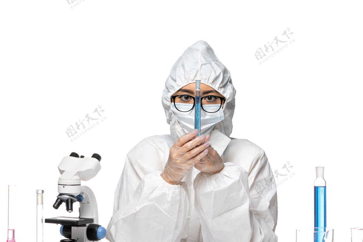 病毒正面图：穿着特殊防护服的年轻女化学家拿着浅白色背景上蓝色溶液的烧瓶——化学病毒实验室特殊烧瓶化学