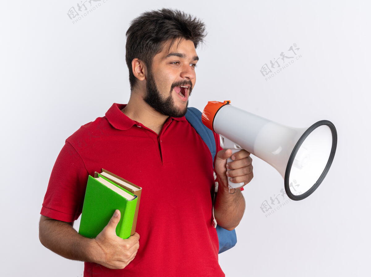 年轻年轻的留着胡子的学生 穿着红色马球衫 背着背包 拿着笔记本 对着扩音器高喊着 站在那里开心又积极笔记本红色喊叫
