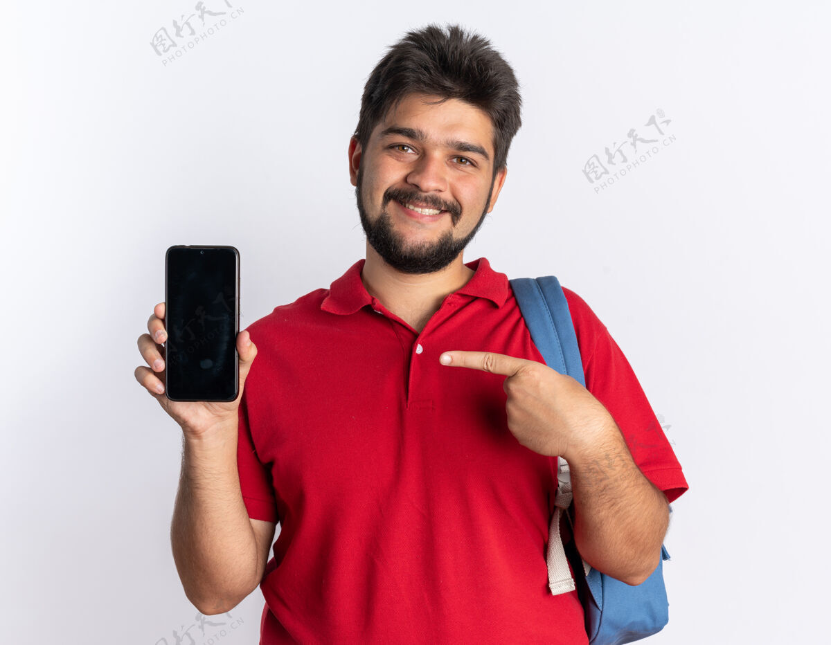 欢呼年轻的留着胡子的学生 穿着红色马球衫 背着背包 拿着智能手机 用idex的手指指着它 站在那里愉快地微笑着小伙子手机学生