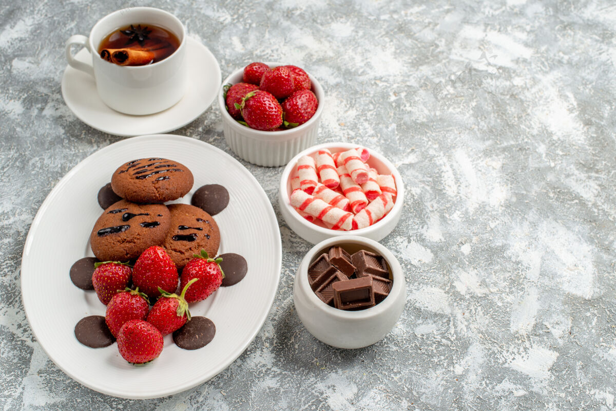 左边底视图饼干草莓和圆形巧克力椭圆形盘子碗糖果草莓巧克力肉桂茴香茶在灰白色的桌子左侧巧克力桌子肉桂