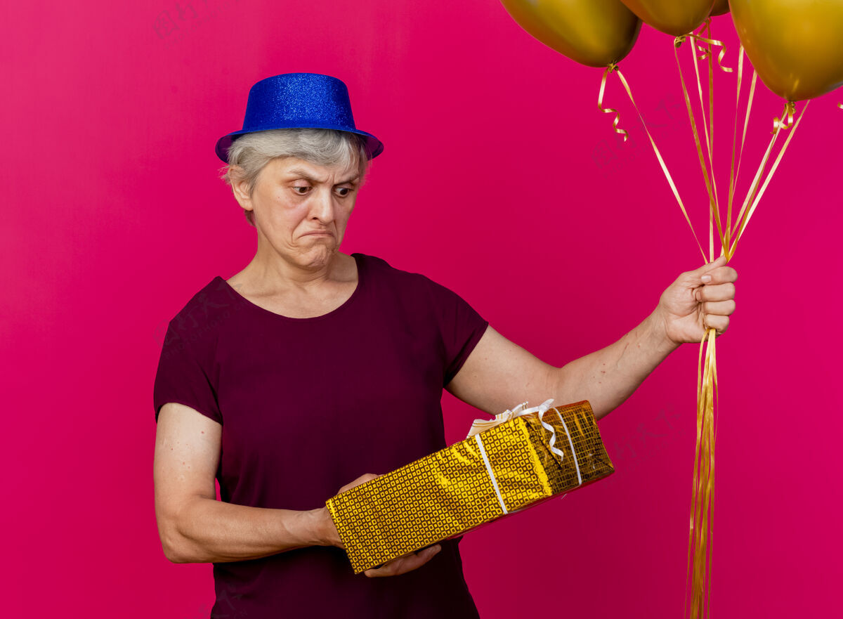 氦一个戴着派对帽的老妇人拿着氦气球 看着粉红色的礼盒女人聚会礼物