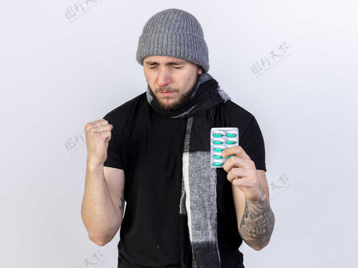 保持戴着冬日帽和围巾的年轻白种人 手上攥着拳头 手里拿着一包白色的药用胶囊拳头疾病冬天